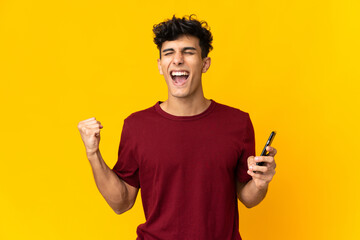 Young Argentinian man isolated on yellow background using mobile phone and doing victory gesture