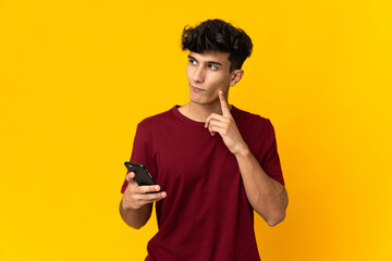 Young Argentinian man isolated on yellow background using mobile phone and thinking