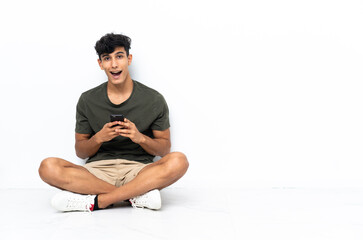 Young Argentinian man sitting on the floor surprised and sending a message