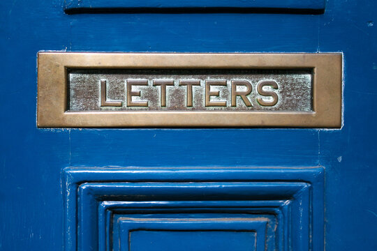 A metal mail slot on a door that says letters.  Mailbox for correspondence.  Wooden door with a metal letter / post slot.