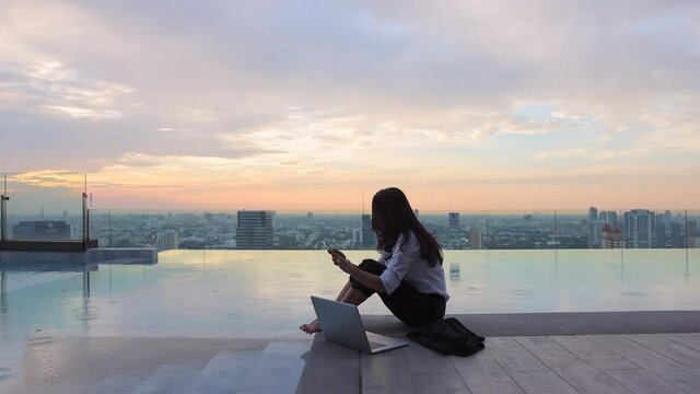 Summer day lifestyle asian woman relax and chill near luxury pool on skyscraper. Asian woman using app on a smartphone and working on a laptop and Blurred background skyscraper, Beautiful sky.