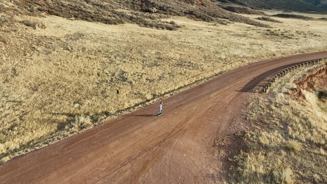 senior male is slowly riding an one-wheeled electric skateboard on a dirt ranch road at Colorado foothills, aerial view of fall or winter scenery