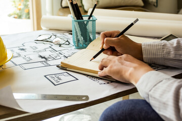 Young engineer writing information on blueprint notebook for workplace meeting, engineer's hand working with tools on a background of project sketches.
