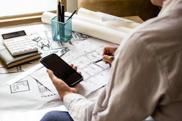 A young engineer compares data between on a smartphone and blueprints for a workplace meeting, an engineer's hand working with tools on a background to draft a project.