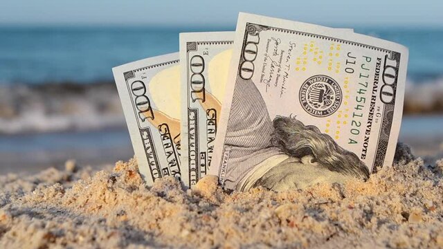 Three one hundred dollar bills half buried in sand on sandy seashore close-up. Three paper dollar bills half in sand on sea beach. Concept money, finance, investment, wealth, poverty, financing, cash