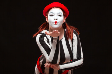 Fototapeta na wymiar Portrait of female mime artist performing, isolated on black background. Woman blows a kiss posing in striped dress