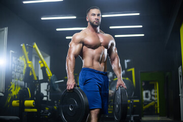 Fototapeta na wymiar Handsome muscular athlete man is standing in a gym holding barbell discs in both hands