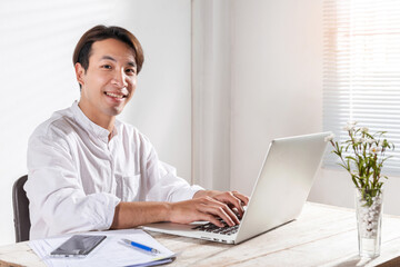 Fototapeta premium A Smart Asian young businessman working at home office pleased happy cheerful on online working or learning using laptop computer getting a new job offer or make good selling. Work win happy concept