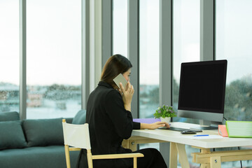 Asian female businesswoman in black suit sitting typing keyboard computer working in living room at home office on call with company corporate colleague talking discussing brainstorming on rainy day