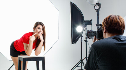 Studio shot Asian pretty female model in red shirt stand smile posing with chair while male photographer using camera shooting picture on backstage with lighting softbox spotlight reflector tripods