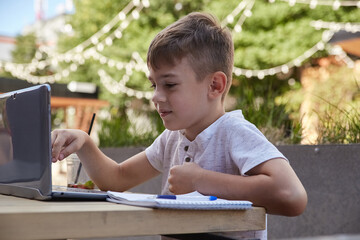 little schoolboy sits outdoors, types on keyboard of laptop. smiling caucasian boy sitting at table at veranda of cafe, studying with computer, doing homework. distant learning, online education