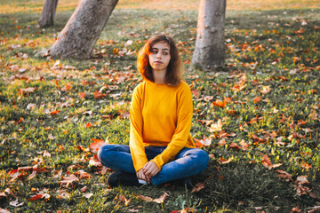 Curly young girl in yellow sweater and jeans sitting on fall grass with dry leaves. Autumn mood