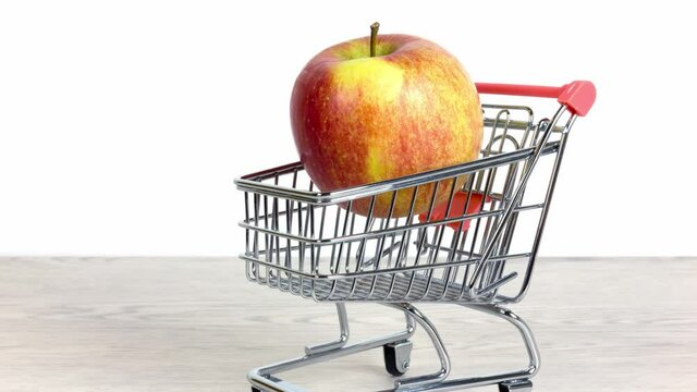 Red and yellow apple inside a miniature shopping cart on a white wooden table over a white background