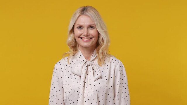Smiling charming elderly gray-haired blonde woman lady 40s years old in pink shirt looking camera wink eye blink isolated on plain yellow background studio portrait. People emotions lifestyle concept