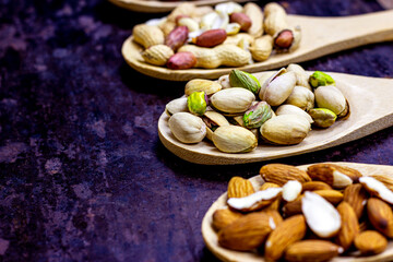 Different nuts: almonds, pistachios, peanuts, hazelnuts in light wooden spoons on black metal background.