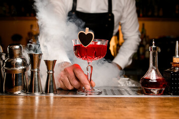bartender accurate holds in his hand a glass of red alcoholic drink decorated with wooden heart