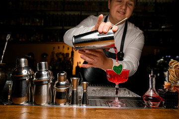 bartender neatly pouring red fresh alcoholic drink into glass decorated with small wooden heart