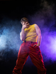 a young muscular man in a bright outfit poses in a pretentious pose, style and grotesque. Attractive guy with pumped-up arms in a mesh T-shirt and red pants, photo in smoke