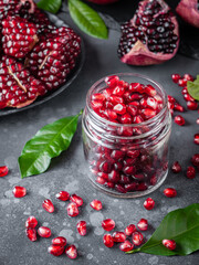 Pomegranate seeds in a jar surrounded by peeled pomegranates and lemon leaves on a gray background