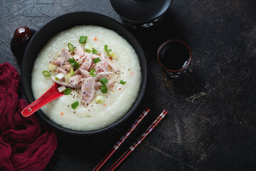 Asian congee served with chicken meat in a black bowl, high angle view on a dark brown stone background, horizontal shot with space
