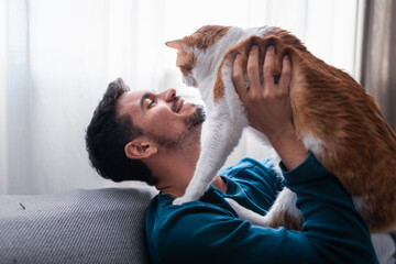 young man sitting on a gray sofa holds in his arms a brown and white cat 