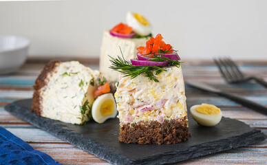 Snack, sliced Swedish salad cake with herring, potatoes, egg and sour cream mousse on black bread on a slate or stone board on a wooden background. Christmas and Easter recipes.
