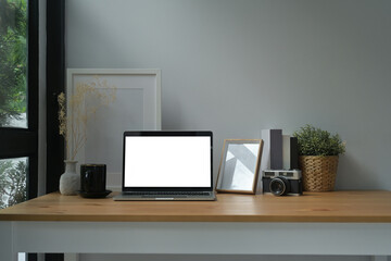 Photo of a comfortable workplace surrounded by a white blank screen computer laptop, empty picture frame, wicker basket, vintage camera, coffee cup, jar and books.