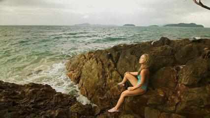 Woman meditates, relaxes on rock crack reef hill in stormy morning cloudy sea. Mother of ocean gives birth to him