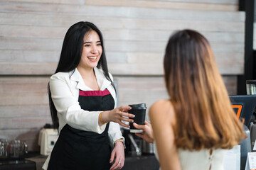 Young pretty caucasian waitress, a small company owner, a barista, and a bartender wearing a black apron serves a customer a coffee cup at the bar counter in a cafe shop