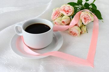 white cup of coffee, beautiful bouquet of white roses and pink satin ribbon heart on white silk background. Congratulations on Valentine's Day, declaration of love