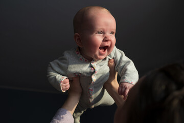 Woman touching foreheads with her newborn child, lifting him up against window at home