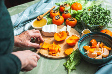 Elderly vegan woman prepares a sweet salad of persimmons, tangerines and soft cheese with honey....