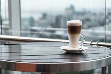 A glass of coffee latte on a table against a blurred background of a cityscape.
