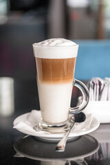 A glass of coffee latte on a table against a blurred background of a cityscape.