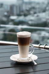 A glass of coffee latte on a table against a blurred background of a cityscape.