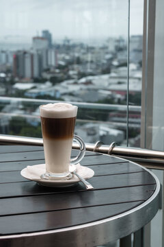 A glass of coffee latte on a table against a blurred background of a cityscape.