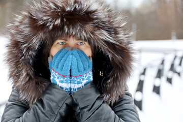 Woman in leather coat with fur hood covers her face by hands in knitted gloves standing on a street during snow. Frost weather in winter