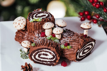 Obraz na płótnie Canvas French dessert called Yule log or bûche de Noël with merengue mushrooms and mint leaves on top of chocolate glazing. Placed in front of Christmas tree. Decorated for Christmas Holidays or New year