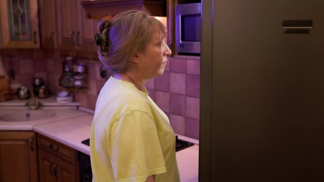 Eating disorder, hungry middle aged woman opens a refrigerator at night and eating unhealthy food.