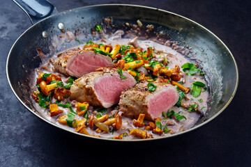 Fried dry aged pork fillet chateaubriand medallion steak natural with chanterelles in walnut cream sauce served as close-up in a classic skillet on black background - obrazy, fototapety, plakaty