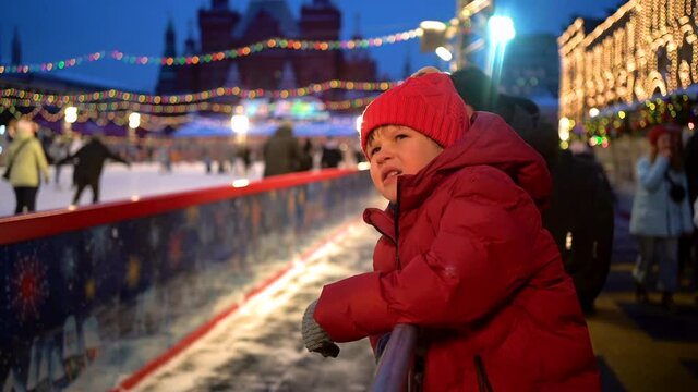 A little boy in a red hat watches people ride on the Christmas ice rink. High quality FullHD footage