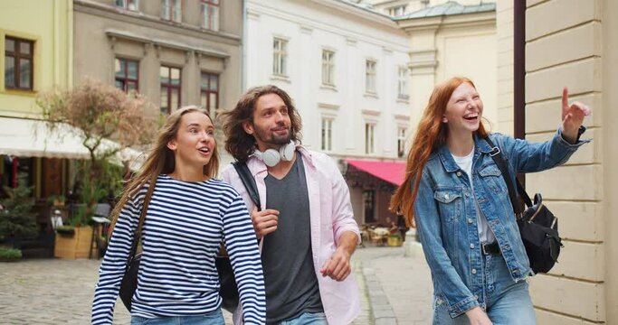 Young female and male exchange students walking on street, having excursion. Best friends going to university, talking and pointing at fascinating buildings. Education, campus, friendship concept.