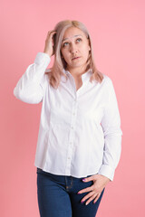 A 40s woman thoughtfully scratches her head while standing on a pink background