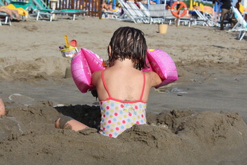 Bambina con i capelli bagnati che gioca in riva al mare con la sabbia.
Indossa un costume da bagno...
