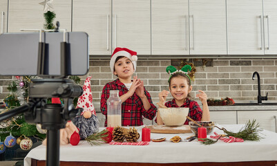 Beautiful children chefs, cute girl and handsome boy wearing Santa hat looking at camera in smartphone mounted on tripod, recording video for vlogging, cooking Christmas bread together in kitchen