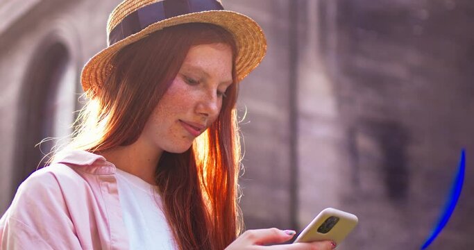 Pretty Caucasian woman with freckles standing near building, using cellphone. Young redheaded girl wearing hat scrolling feed, searching through Internet on smartphone. Social media, technology.