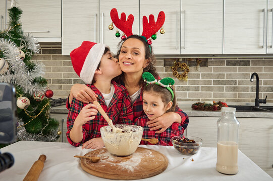 Happy Mother Hugs Her Adorable Kids, All Dressed In Red And Green Plaid Clothes. Cute Son, Young Teenager Kissing Mom On Cheek While Cooking Together And Recording Video For Christmas Food Video Blog