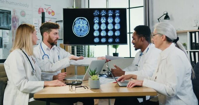 Good-looking confident experienced hardworking multicultural doctors sitting around boardroom table in modern clinic and get aquainted with report,slow motion