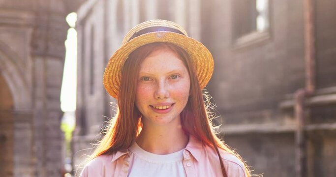 Portrait of good-looking female girl wearing hat standing on street on sunny summer day. Young redheaded woamn with freckles looking to camera ans smilling sincerely outdoors. People, beauty concept.