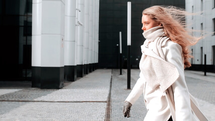 A girl in a white coat and a scarf is walking down the street, smiling. Business center in the...